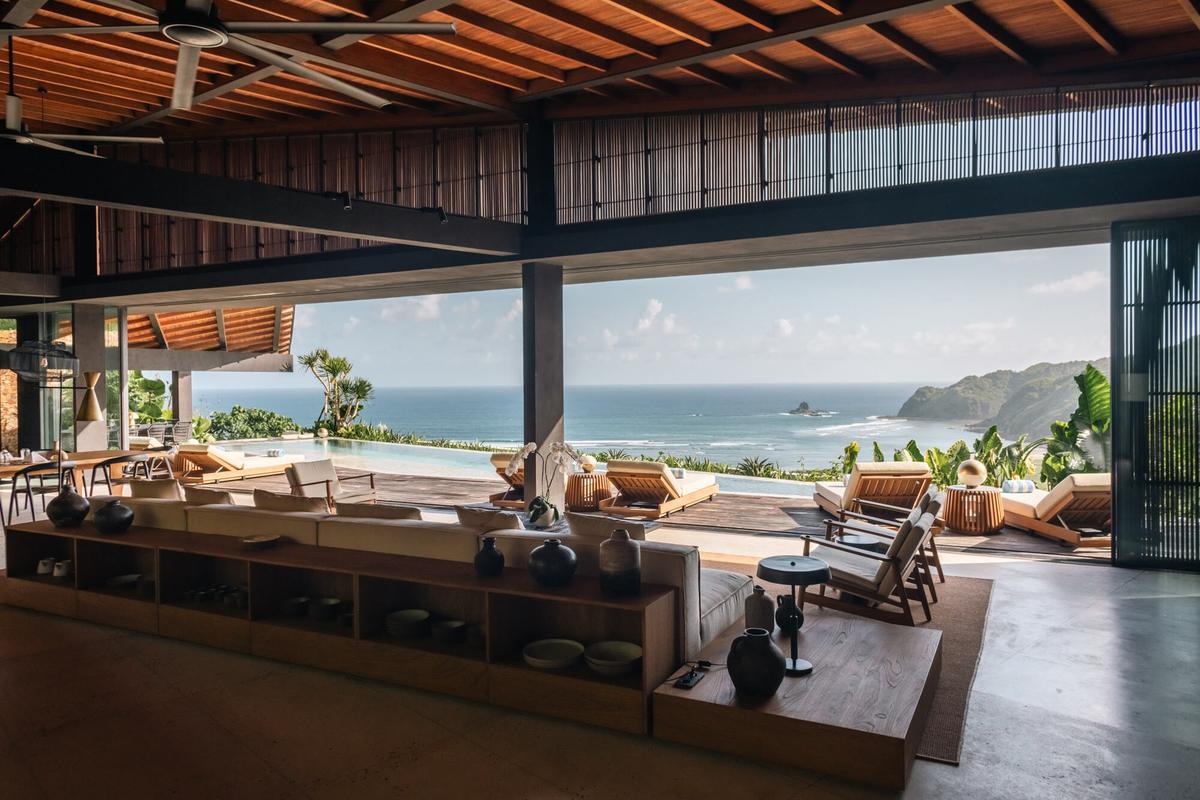 An open living room under a timber roof, looking across a pool to the bay